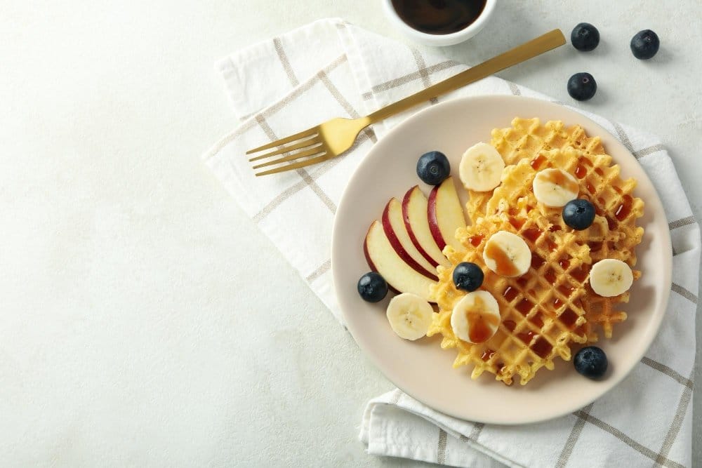 Plate of golden waffles topped with banana slices, blueberries, and syrup, served with apple slices and a cup of coffee in a bright setting.