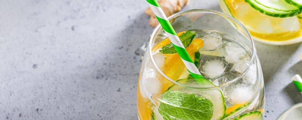 Hugo Spritz Recipe – A Refreshing Summer Cocktail 3 A close-up of a Hugo Spritz cocktail garnished with fresh mint, cucumber, orange slices, and served with a green-striped straw on a light gray surface.