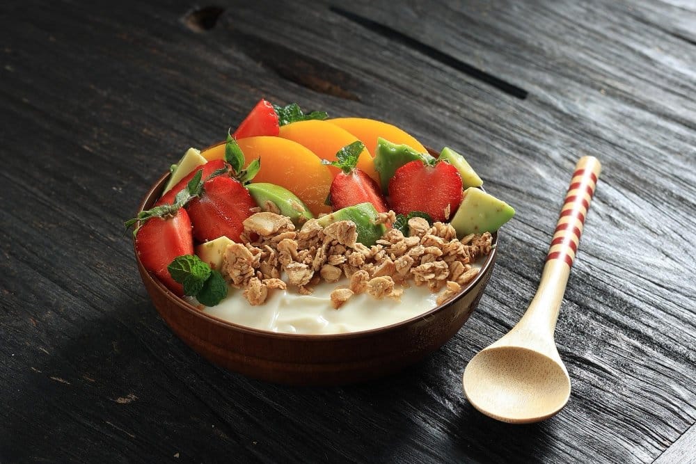 What is the Healthiest Breakfast You Can Eat? 2 Healthy yogurt bowl with fresh fruits like strawberries, peaches, and avocado, topped with granola and mint leaves.