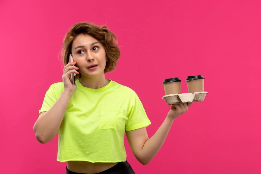 Can I Drink Starbucks and Lose Weight? Best Low-Calorie Tips! 2 Can I drink Starbucks and still lose weight: Woman in a neon green shirt holding coffee cups and talking on the phone against a pink background.