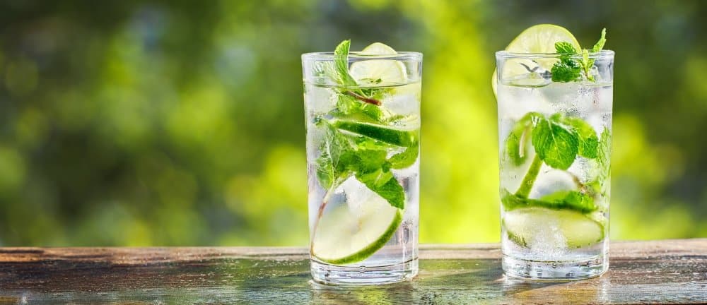 How to Make the Perfect Hugo Spritz Recipe 2 How to Make the Perfect Hugo Spritz Recipe: Two glasses of Hugo Spritz cocktail garnished with fresh mint and lime slices, placed on a wooden table with a blurred green background.
