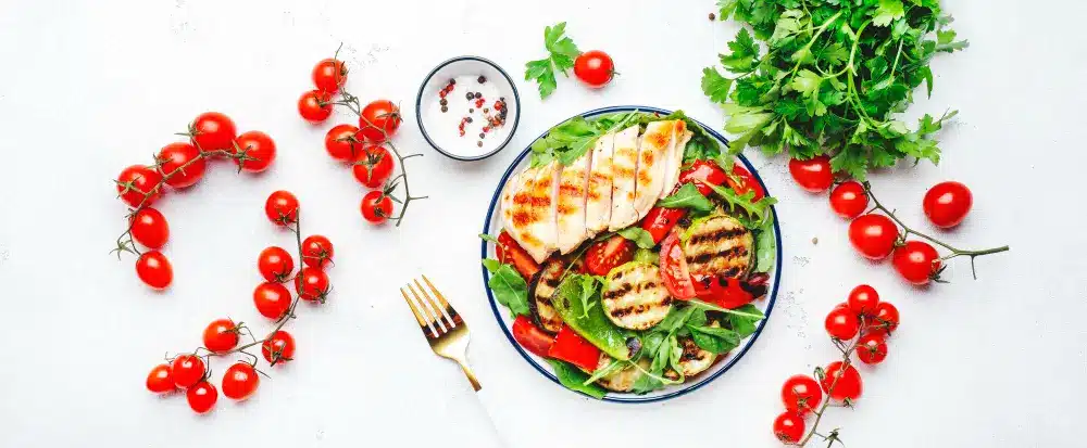 Gluten-Free Meals: A healthy gluten-free meal on a white table featuring grilled chicken, fresh cherry tomatoes, mixed greens, grilled vegetables, and parsley.